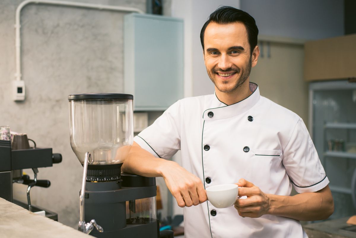 A staff member in chef whites standing at a café counter beside a coffee grinder